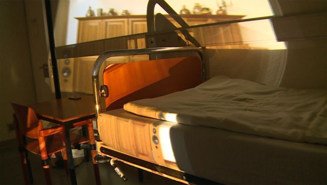 Ein Krankenhausbett, daneben ein Holztisch und -stuhl, angeschienen durch eine Projektion von einem Sideboard