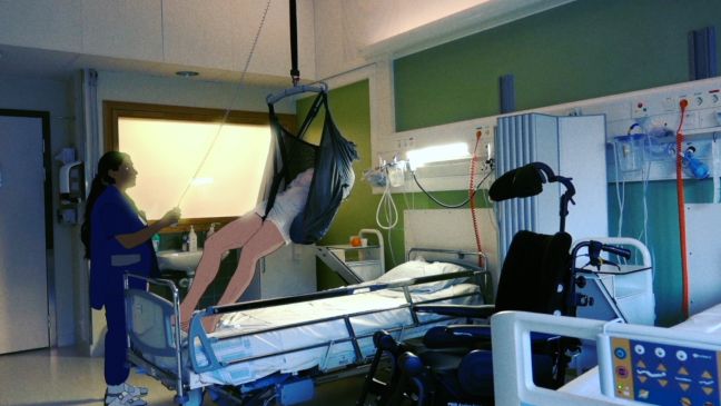ein Mensch wird mit Hilfe einer Trage von einer Krankenschwester aus dem Bett gehoben