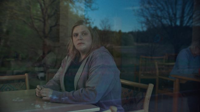 Lea sitzt am Tisch, man sieht sie durch eine Fensterscheibe