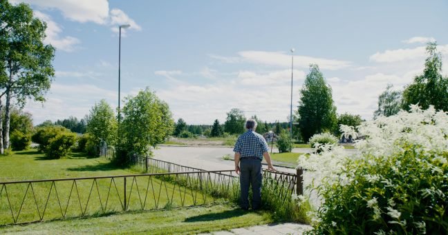 Ein älterer 
Mann steht an einem sonnigen Tag in einem Vorgarten, den Rücken zugewandt