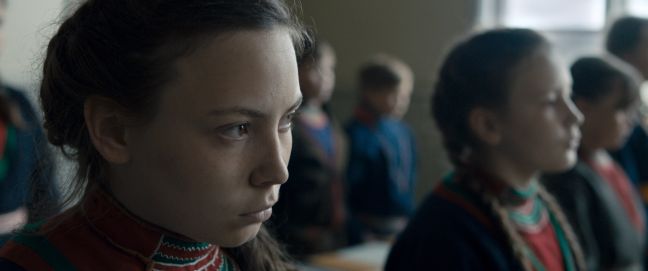 Nahaufnahme von Elle-Marja (Lene Cecilia Sparrok). Sie ist im Klassenraum mit anderen Sámi-Kindern, alle tragen Gákti.