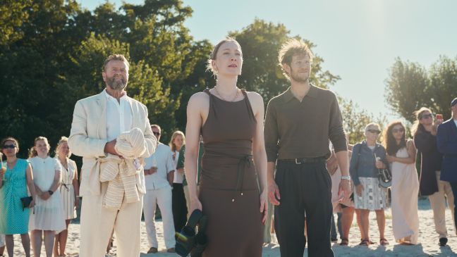Emilie und Karl stehen am Strand und schauen aufs Wasser, die Gäste hinter ihnen