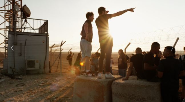 Eine Gruppe Menschen an einem Grenzzaun, ein Mann zeigt in die Ferne über den Zaun