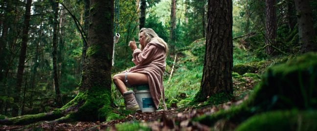 Agnes sitzt auf einem selbstgebastelten Klo im Wald und zündet sich eine Zigarette an
