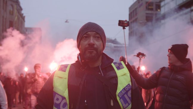 Dialogpolizist Jimmy vor einer Gruppe Demonstranten, im Hintergrund Rauch und angezündete Signalmittel
