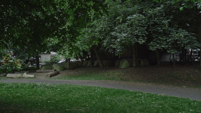 ein Park, unter Bäumen einige grüne Zelte