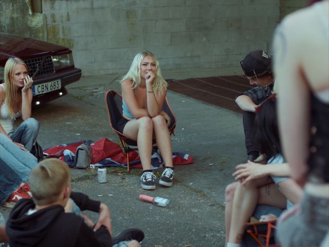 Bella und andere Jugendliche sitzen auf einem Parkplatz und spielen Flaschendrehen