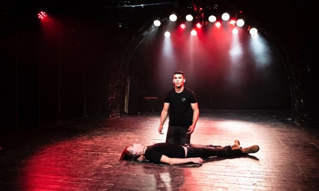 Zwei Personen auf einer Theaterbühne, eine liegt auf dem Boden, die andere kniet neben ihr